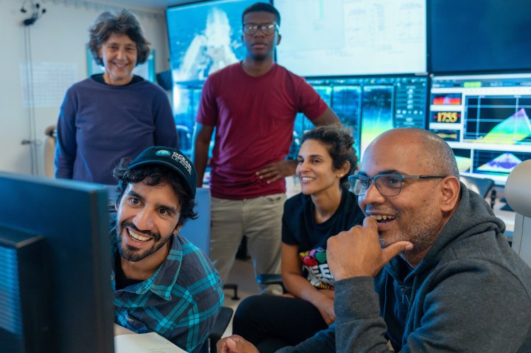 A group of five people in a tech lab, smiling and engaged with computer screens displaying video data from the DORIS lander.